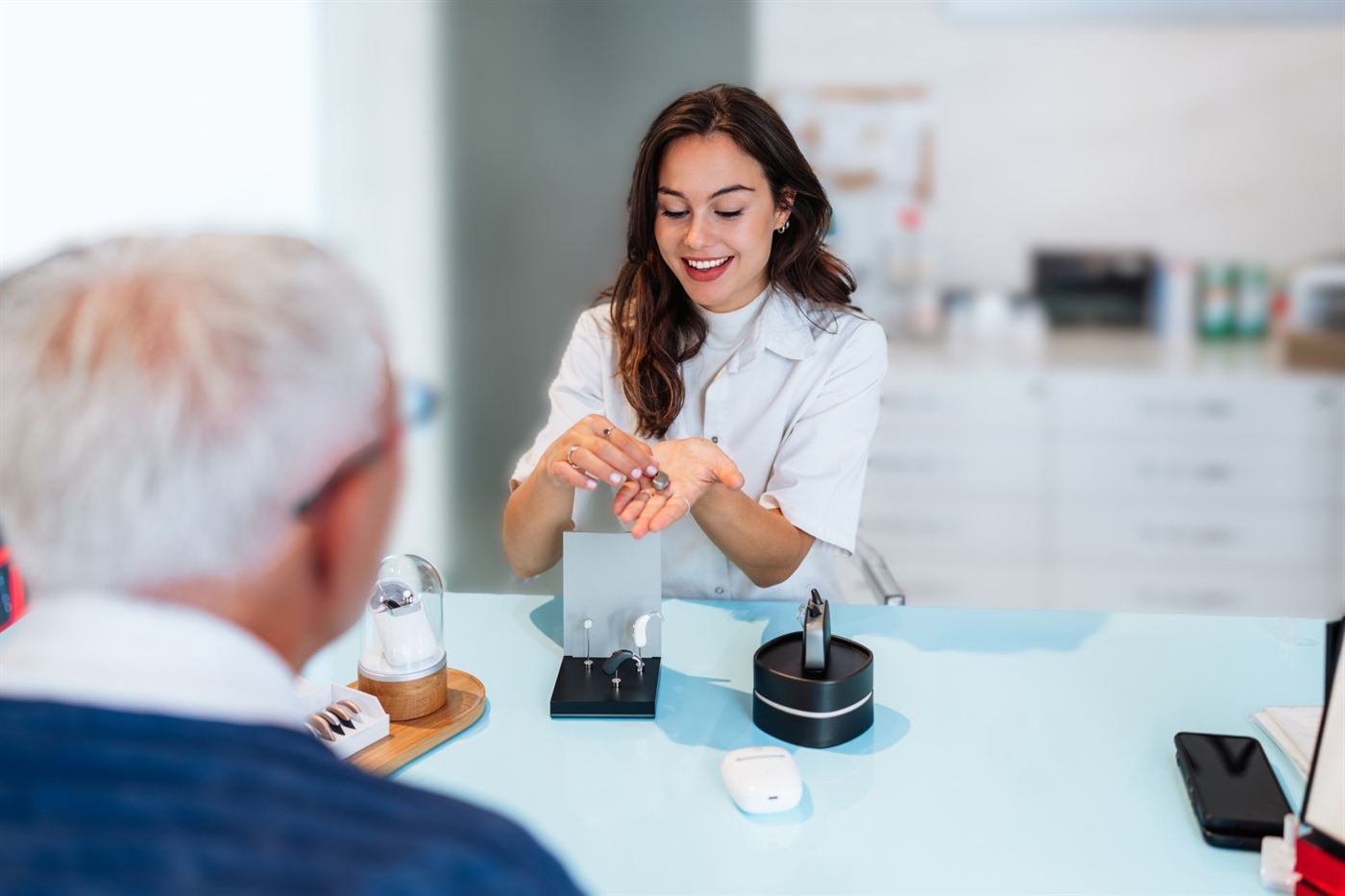 A smiling hearing professional holds a prescription hearing aid while showing hearing aid options to an older patient